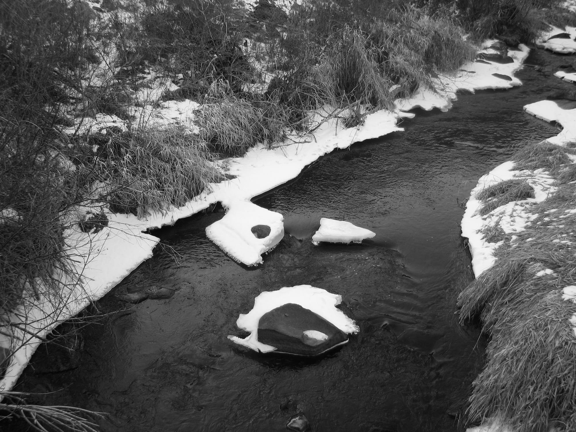 A black and white photo of a creek in winter, with snow on the banks and on rocks