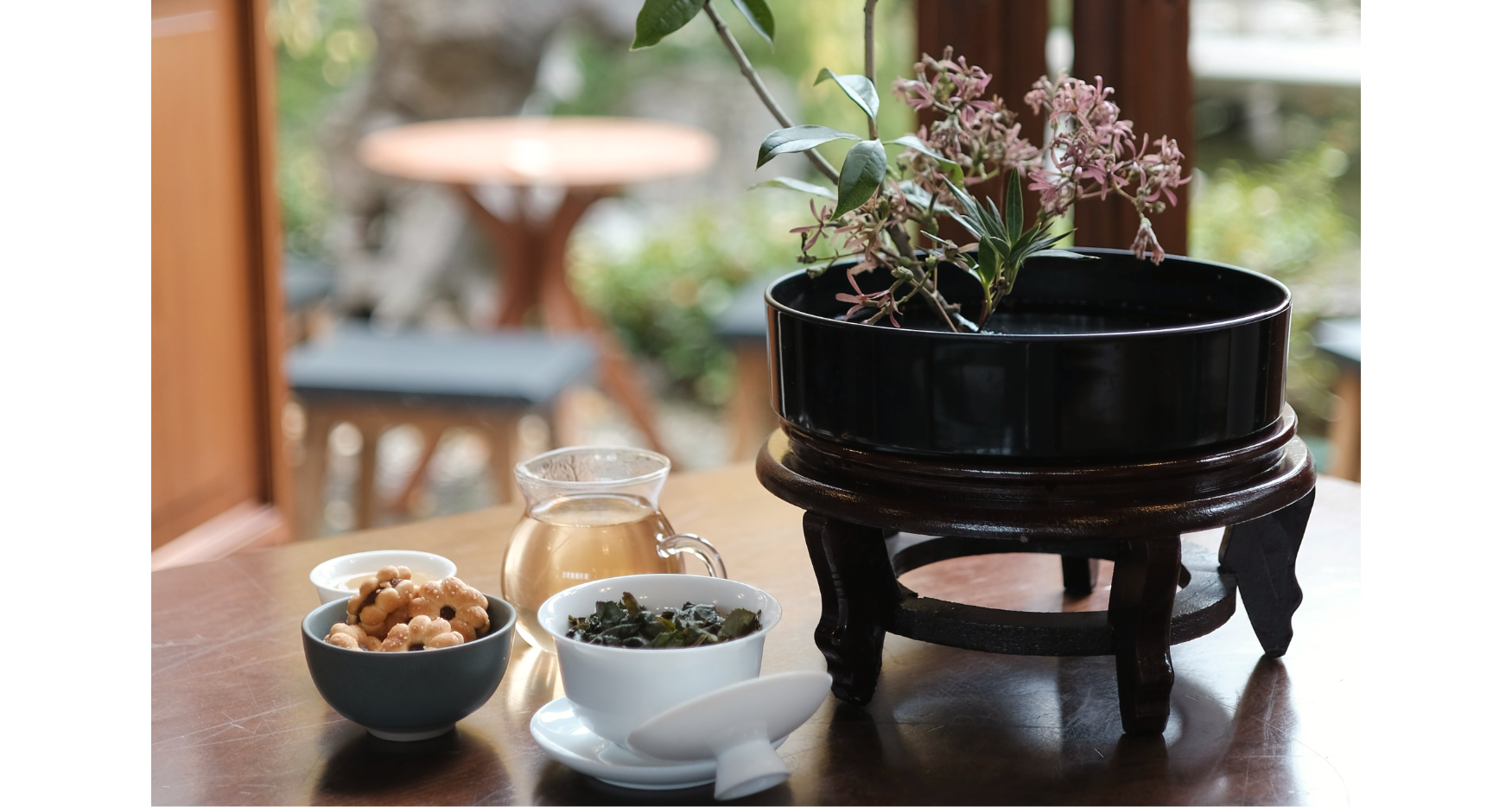 Close-up photo of small bowls and glass teapot, with flowers in a black flat circular vase on a wooden table