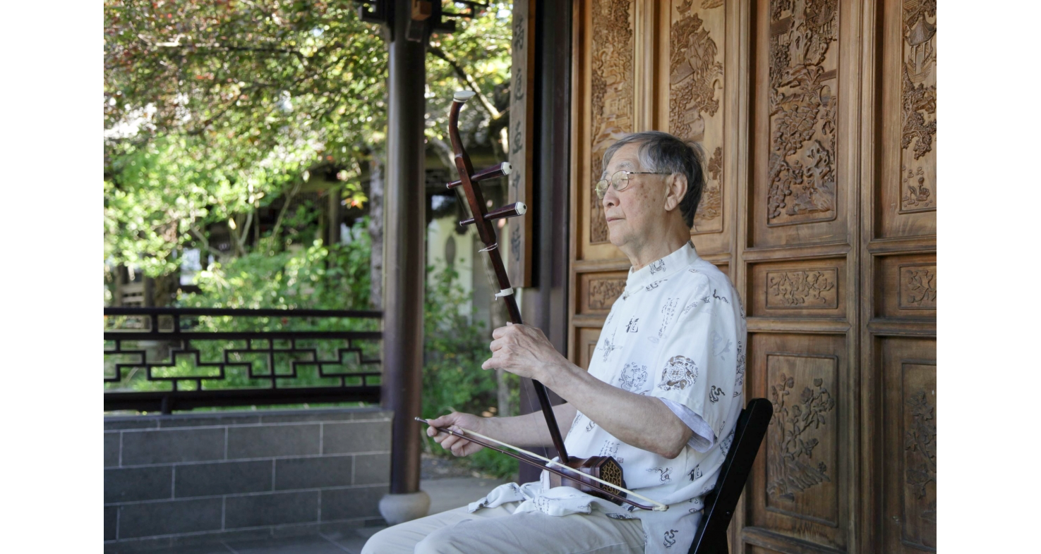 Photo of an older Chinese man in white patterned shirt and glasses, playing the erhu outside under a pavilion in front of a carved screen