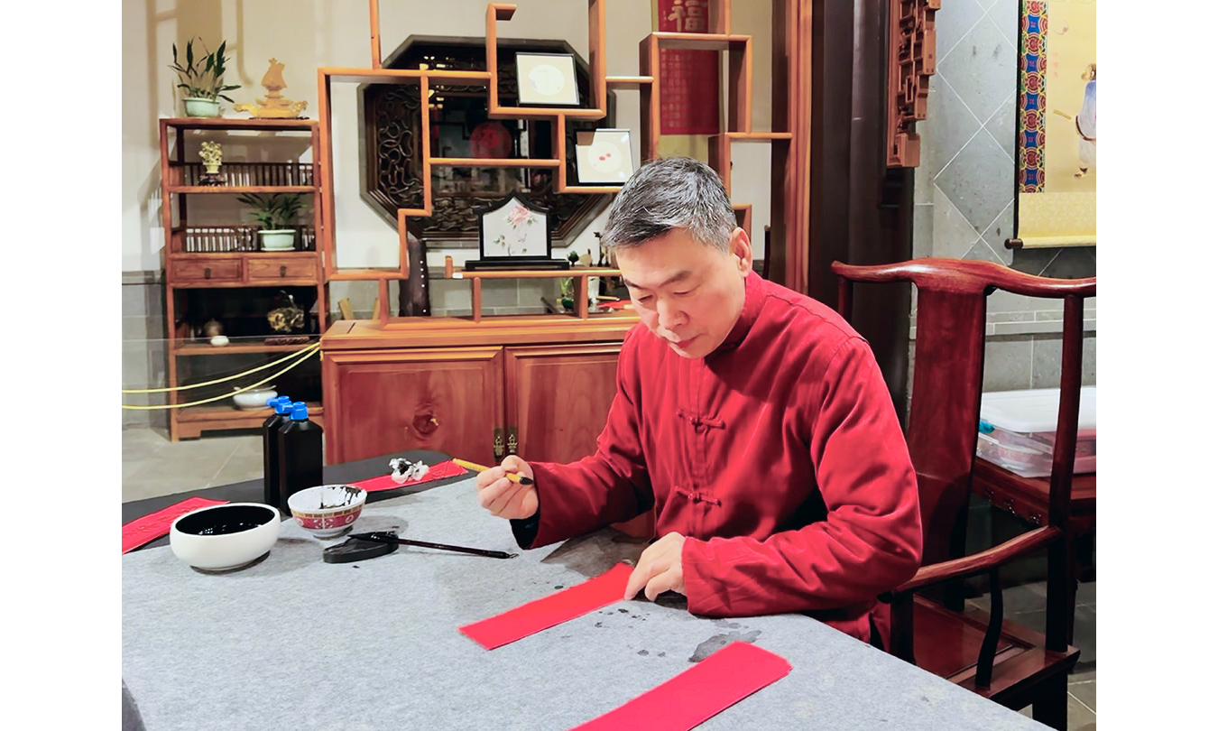 Photo of Chinese man in a in red shirt sitting a table with pen in right hand about to write on a vertical strip of red paper