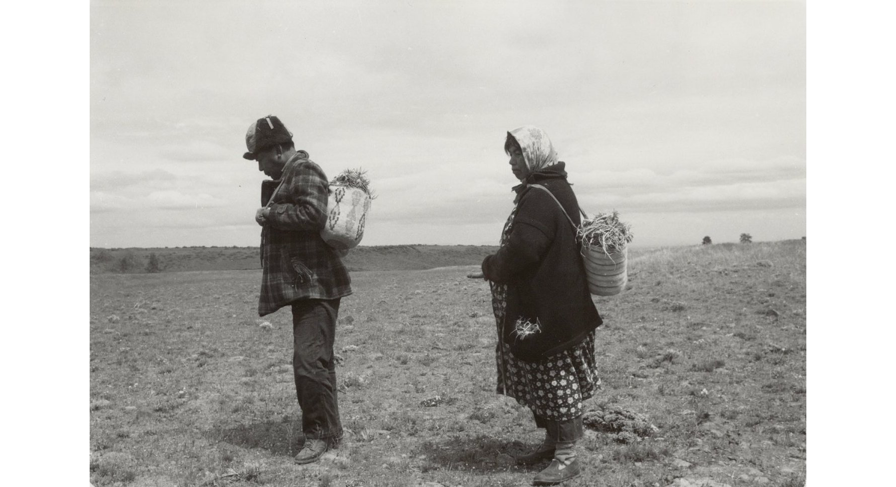 Photo of man in jacket and hat, and women in kerchief and coat, facing left, and carrying filled baskets, on barren meadow
