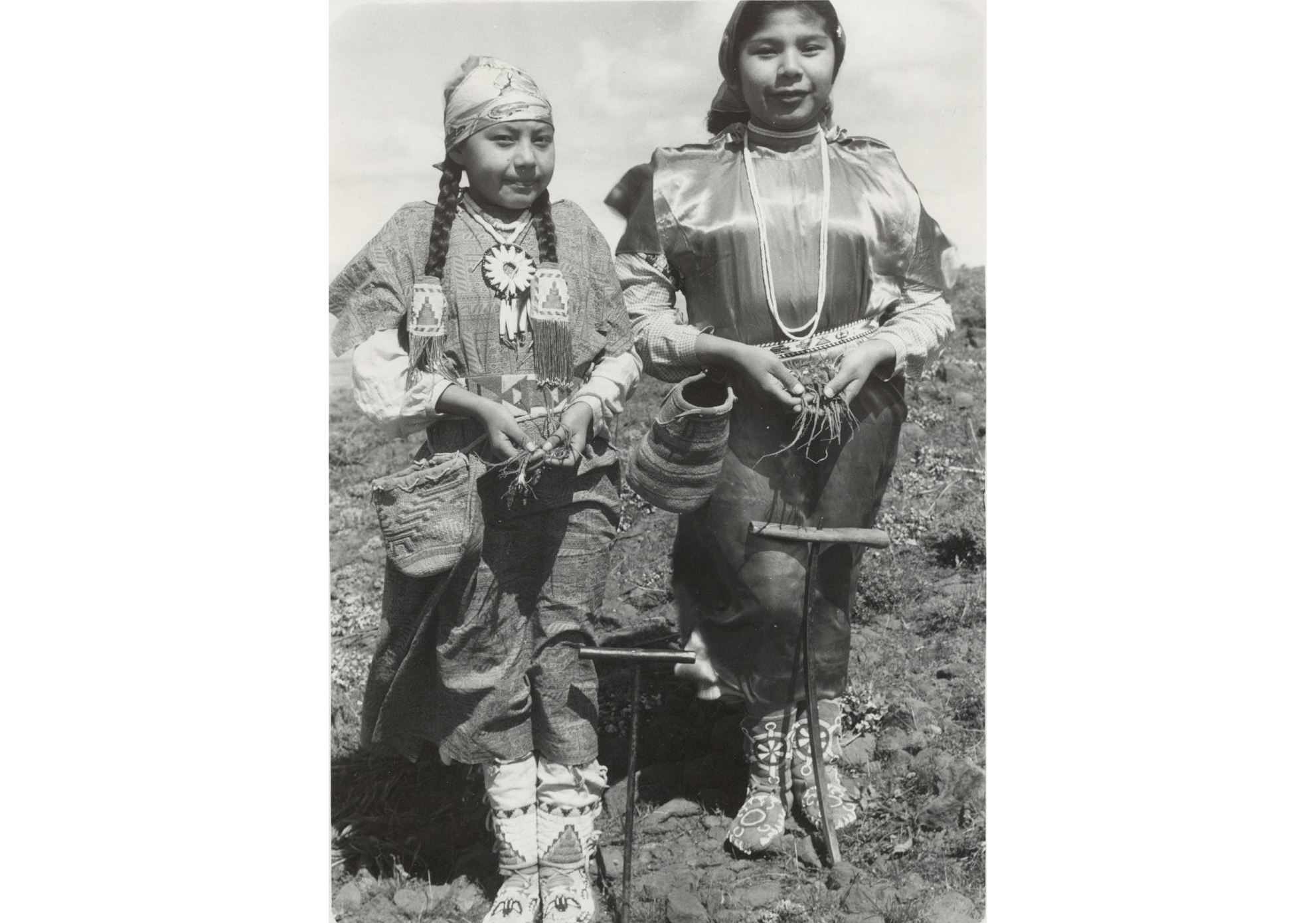 Photo of two Yakama girls in traditional dress with hands clasped in front of them holding roots