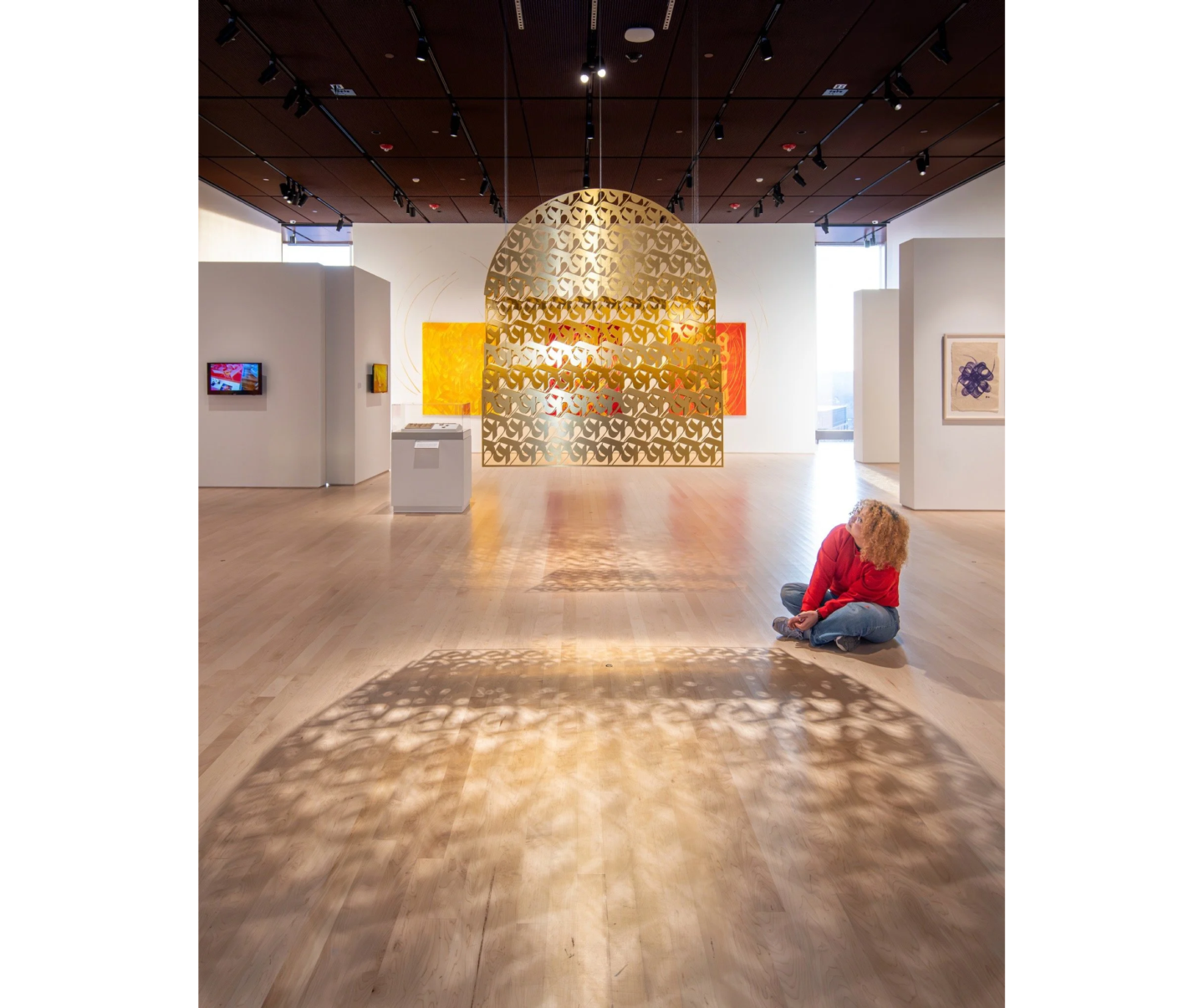 Photo of woman in museum gallery room, seated on wooden floor to right, looking up at golden filigreed screen handing from ceiling