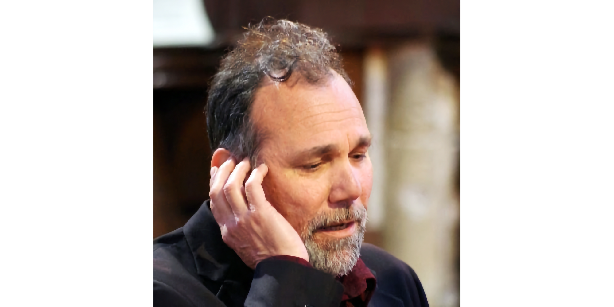 Photo of head of man with mustached and goateed singing, looking to lower right of frame, with his right hand cupped near his right ear