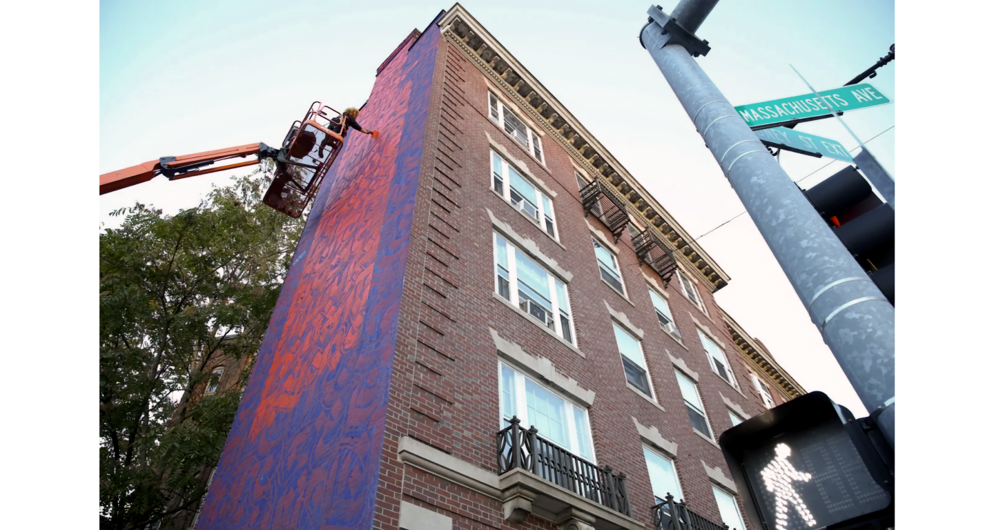 Photo of woman on boom lift painting side of building in bright colors, with brick facade angled in front