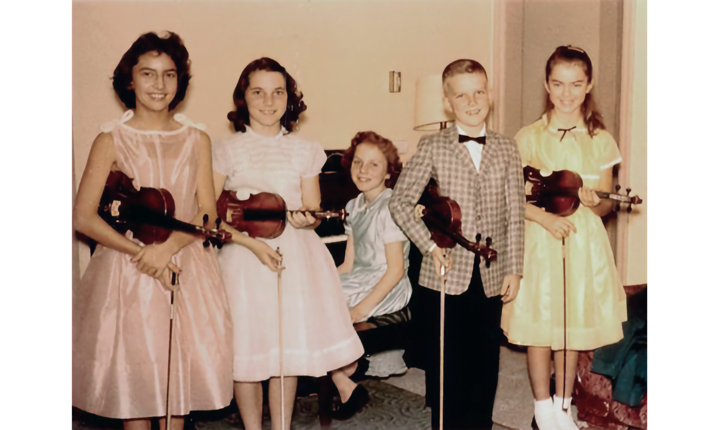 Vintage photo of 5 children, 4 standing with violins, and one seated at a piano behind them.