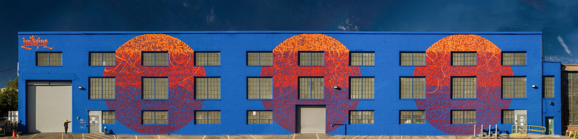 Panorama photo of same woman in lower left corner in front of entire 3-story building with 3 large designs painted in bright colors