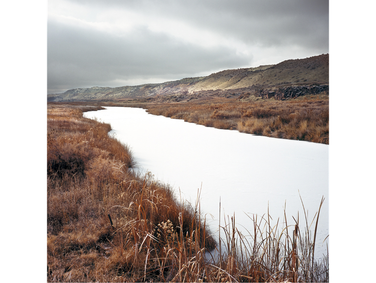 A wno-covered creek runs top left to bottom right, with brown grass on each bank and hills in the backgroun.