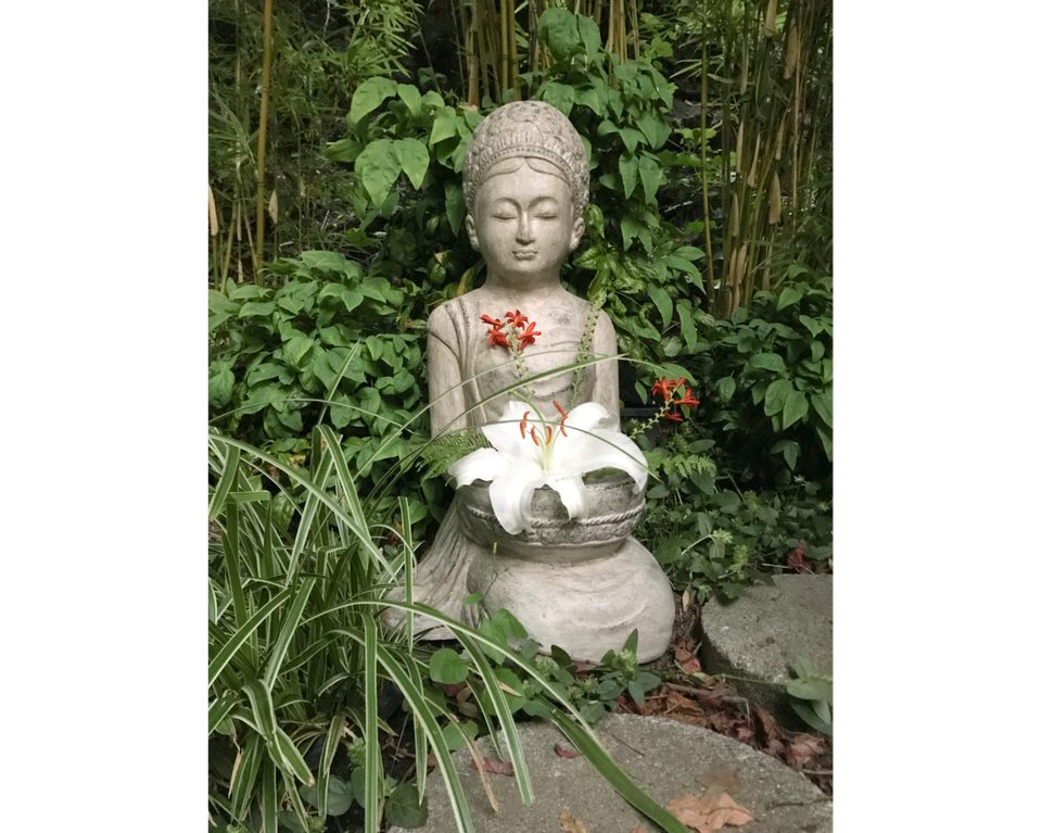 Photo of concrete cast sculpture of Buddhist goddess holding white lily in her hands in front, with ivy and fence in background